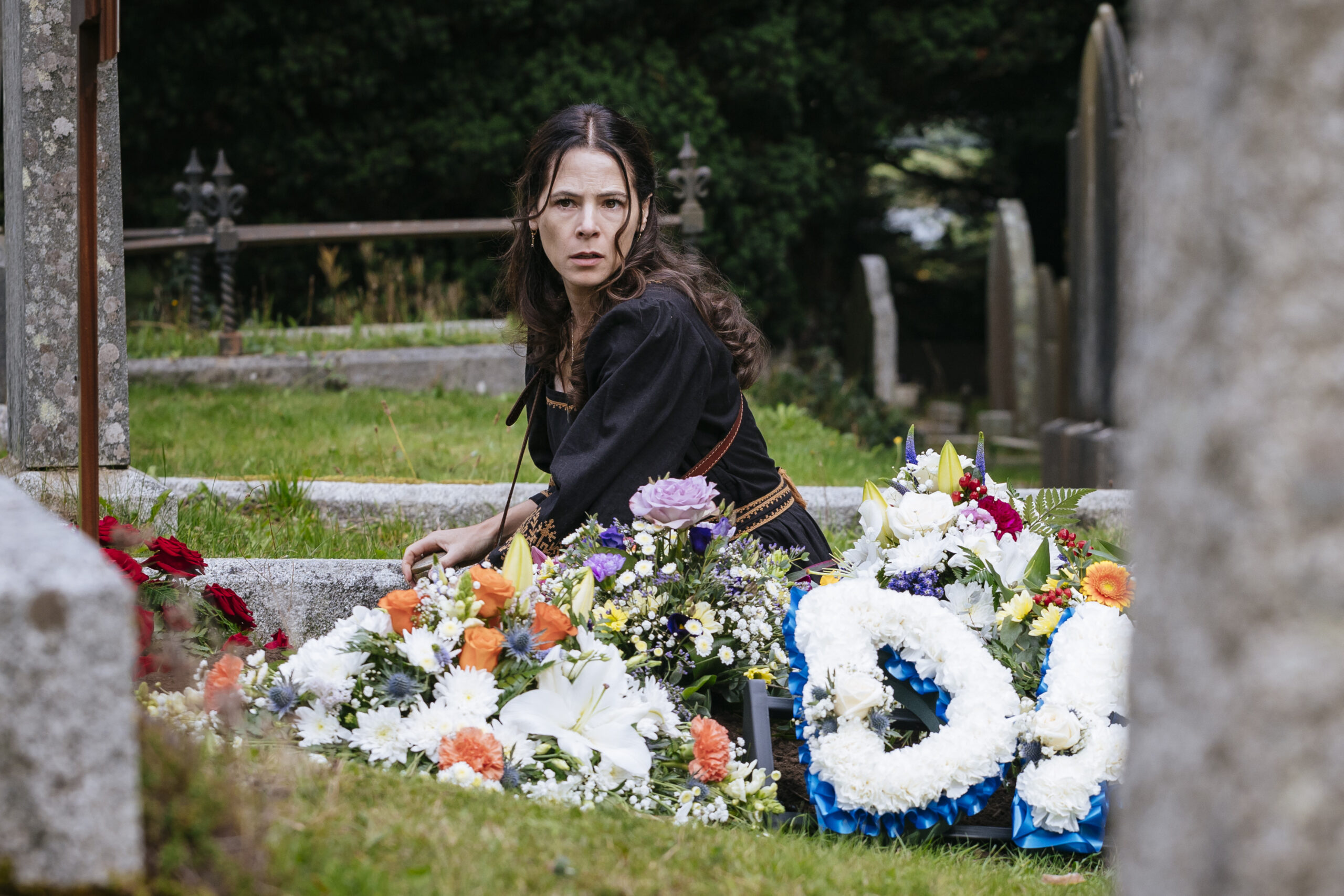 A woman in black sits on the ground in front of a memorial full of flowers